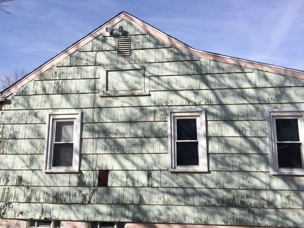Side view of a weathered light green house with three windows and an attic vent under a bright sky