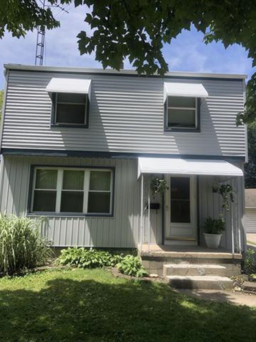 Two-story house with gray siding, white window awnings, and a small front porch with steps