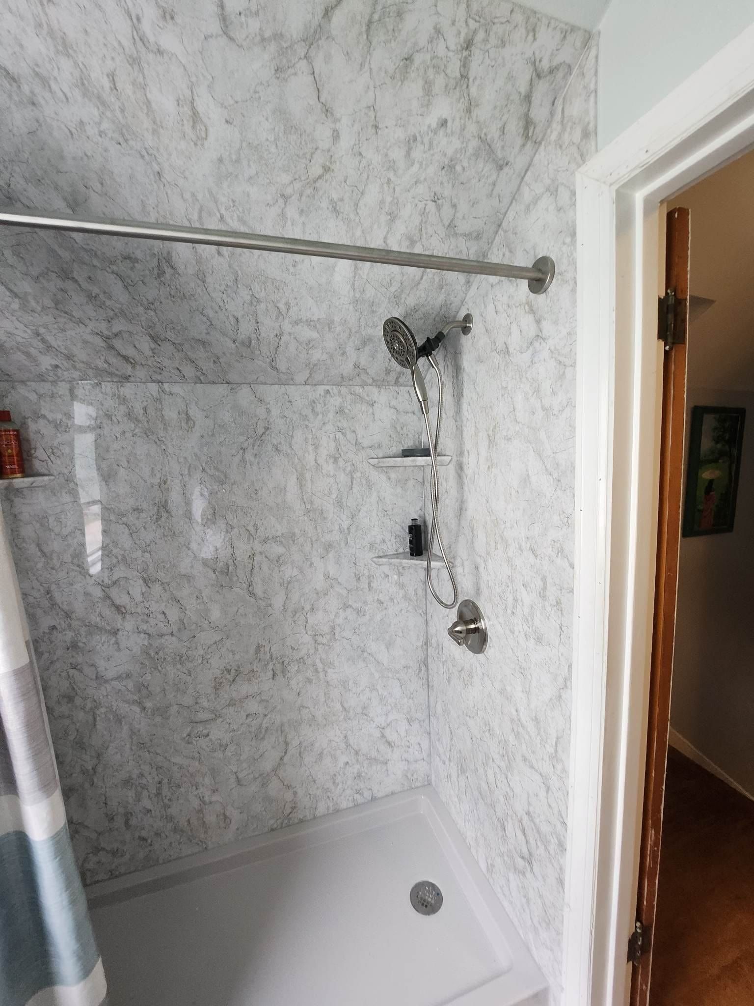 Shower stall with marble-patterned walls, showerhead, and a shelf unit, next to an open doorway.