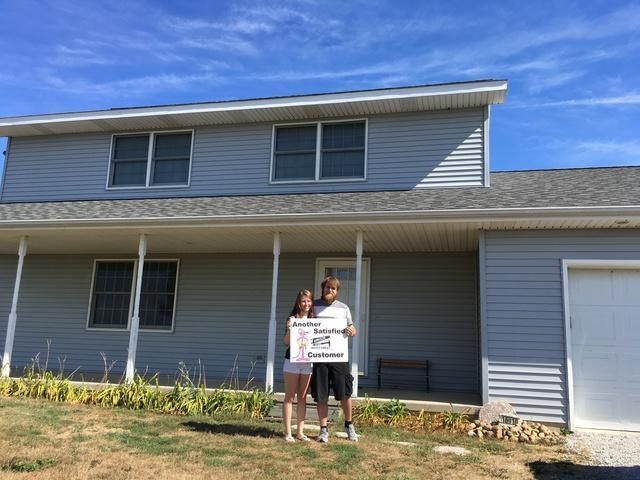 Couple holding a sign in front of a gray two-story house with a porch and attached garage on a sunny day.