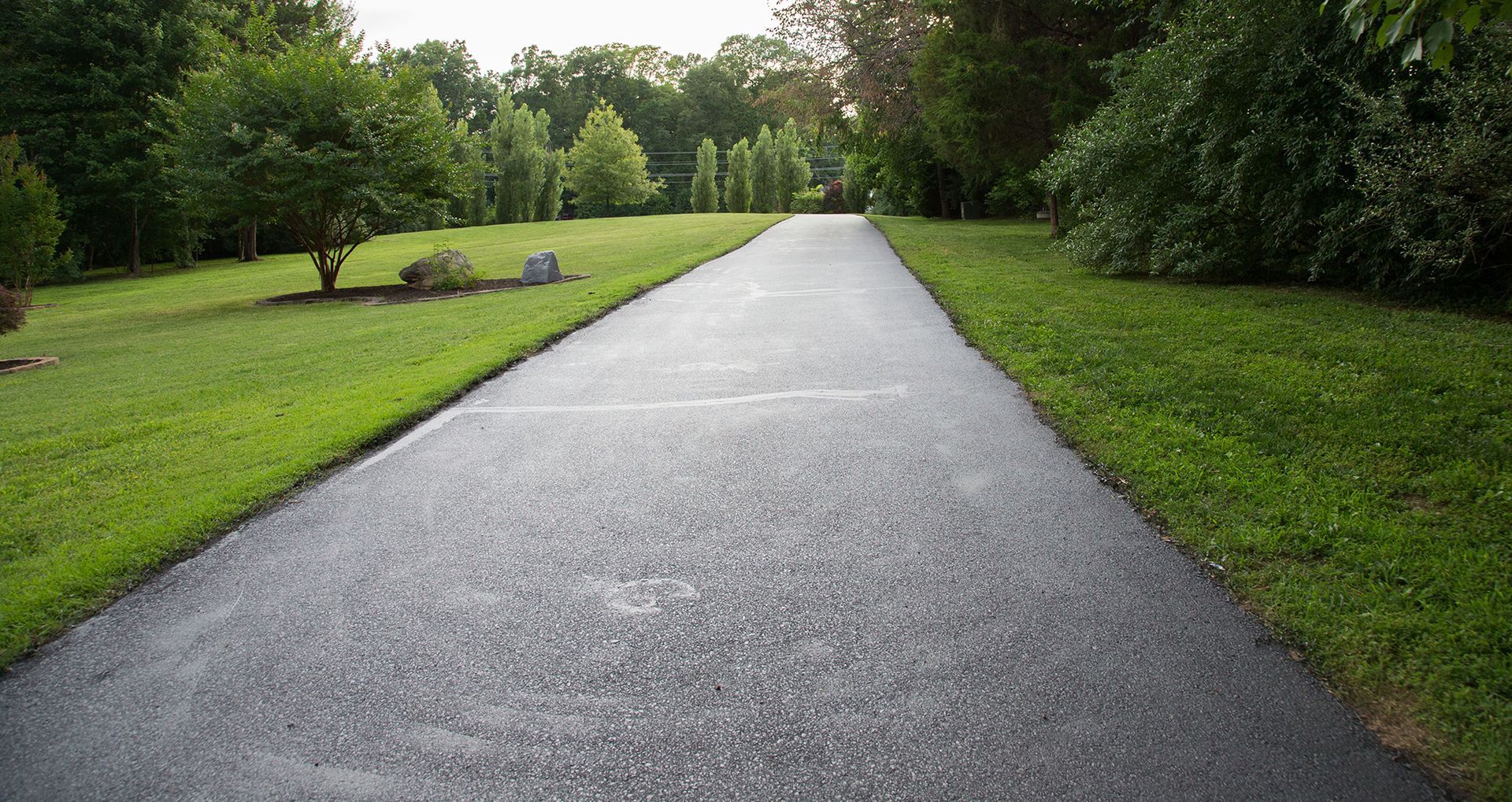 A paved driveway stretches through a grassy lawn surrounded by trees under a bright sky.
