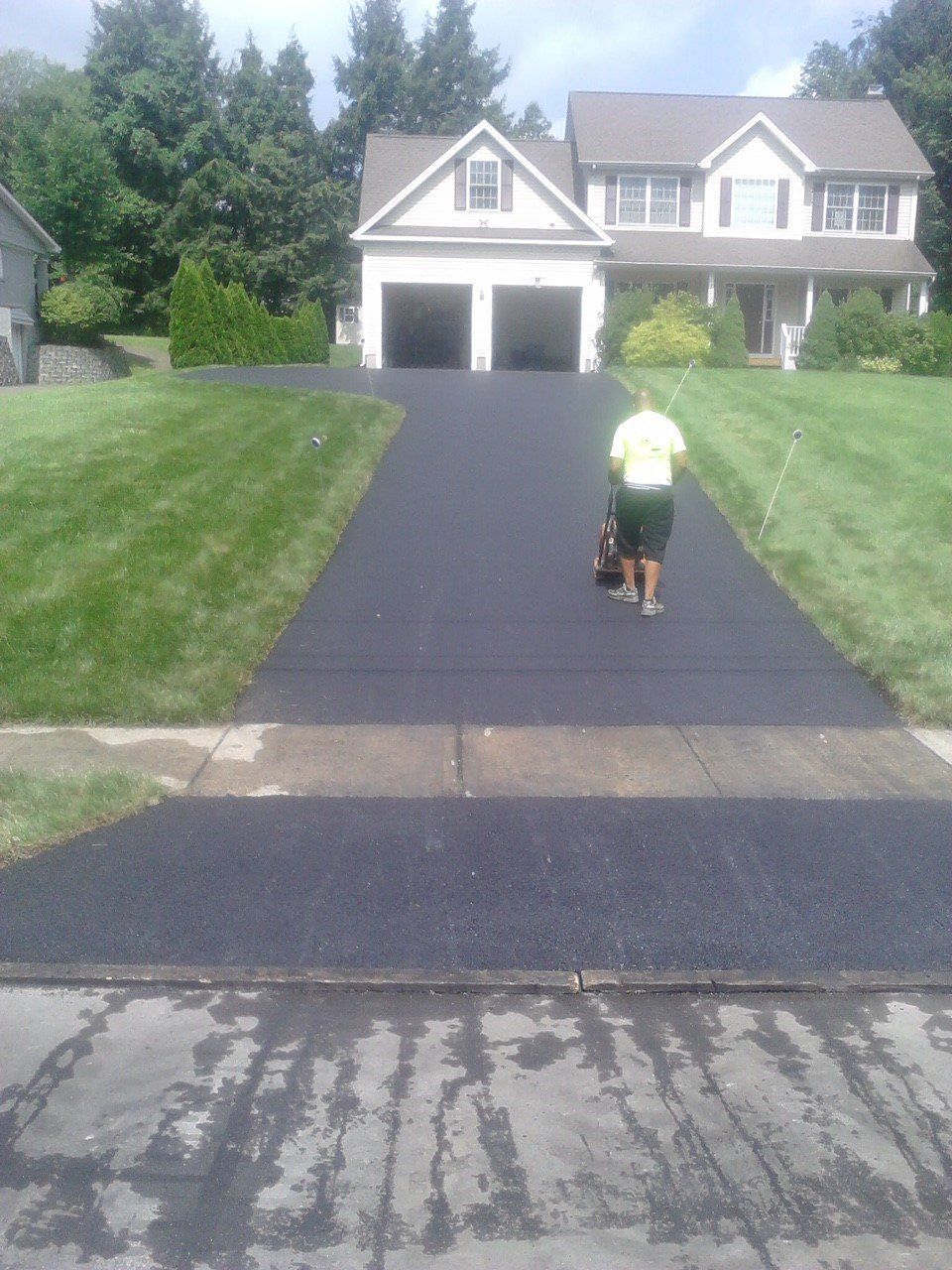 A person in a yellow shirt operates a machine to seal a newly paved driveway leading to a two-story house.