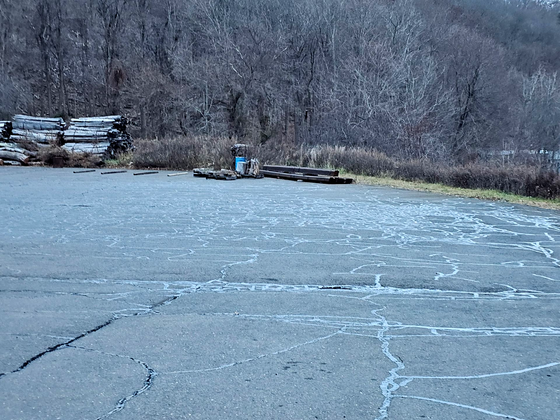 A paved lot with cracks, featuring stacked logs on the left and a small blue structure in the background against woods.