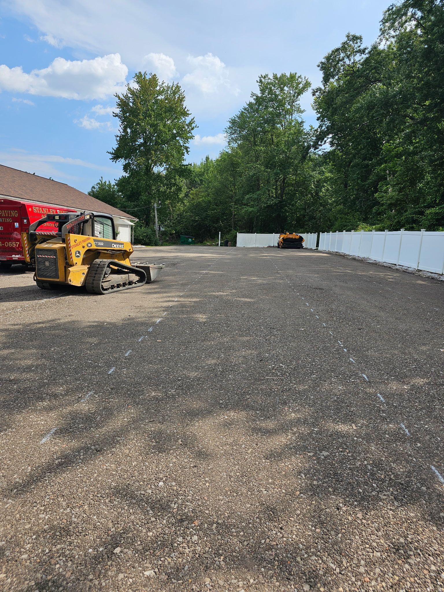 A yellow skid steer sits on a gravel lot next to a red building and a white fence under a partly cloudy sky.