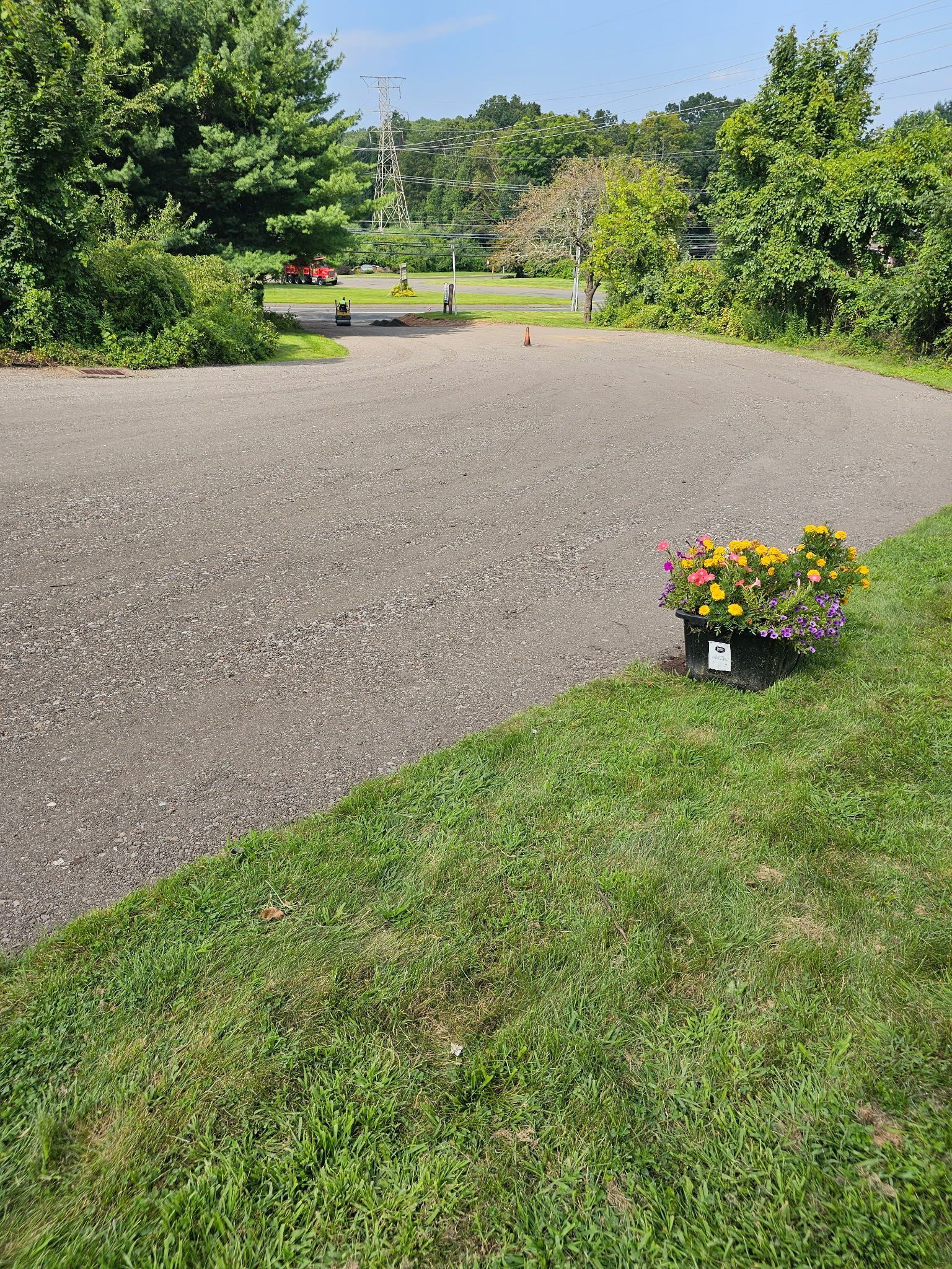 A gravel driveway leads toward a grassy area with trees, featuring a potted flower arrangement in the foreground.