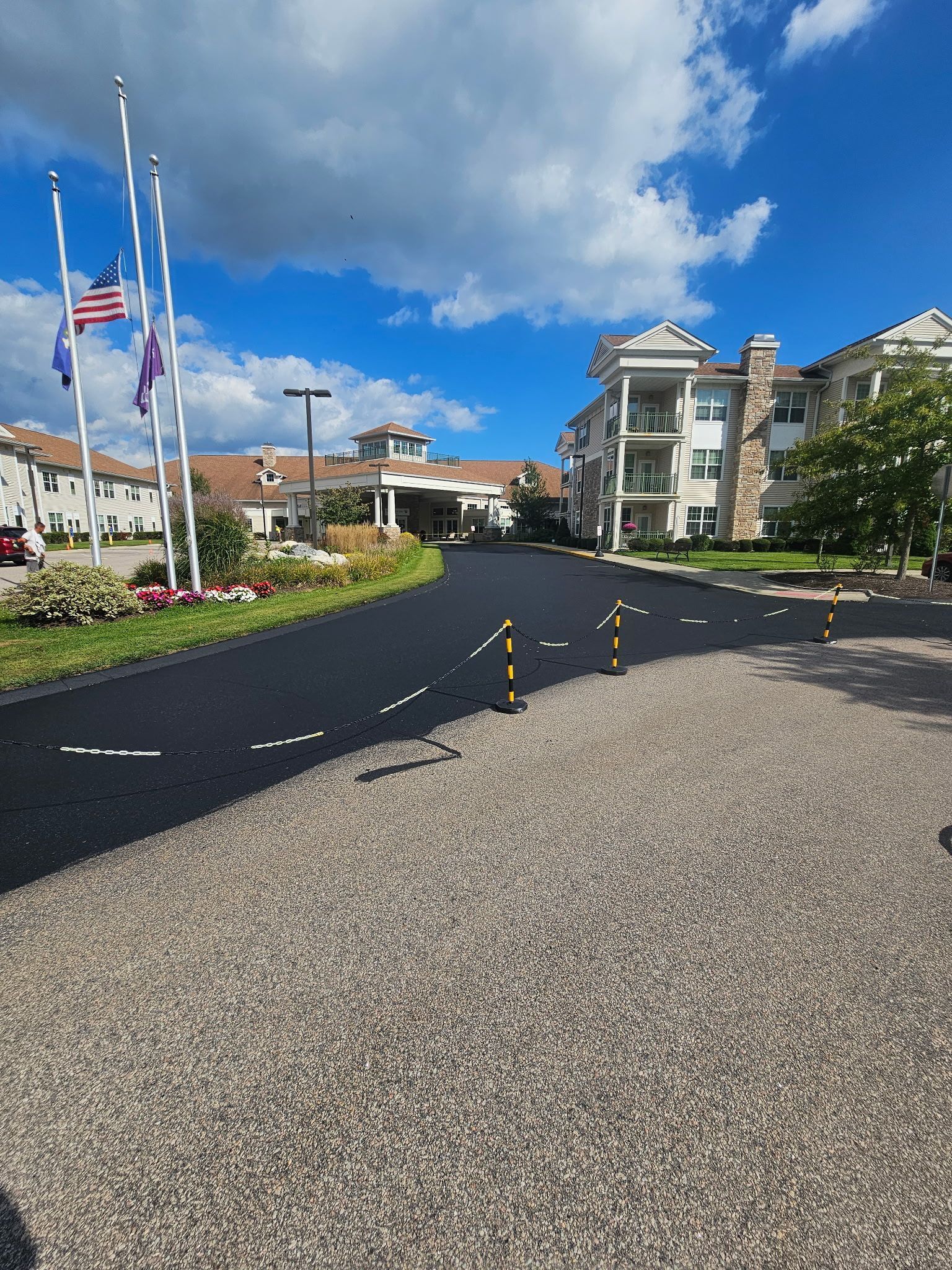 A newly paved black asphalt road leads toward a large, multi-story building under a blue sky with flags in the foreground.