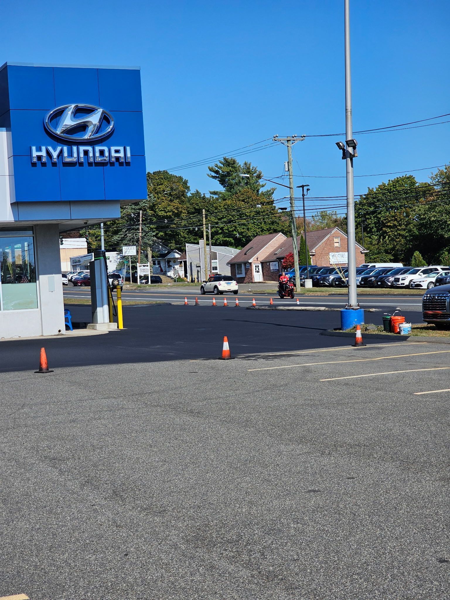 A Hyundai dealership lot with newly paved black asphalt, orange traffic cones, and trees under a clear blue sky.