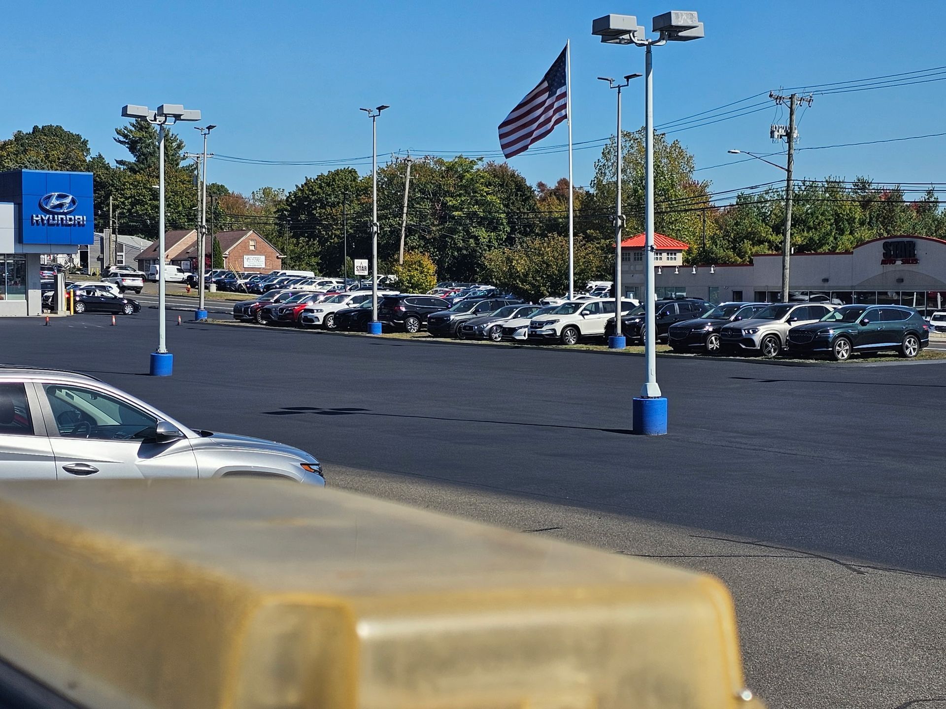 A car dealership lot under a clear blue sky, featuring a Hyundai sign, parked vehicles, and an American flag on a pole.