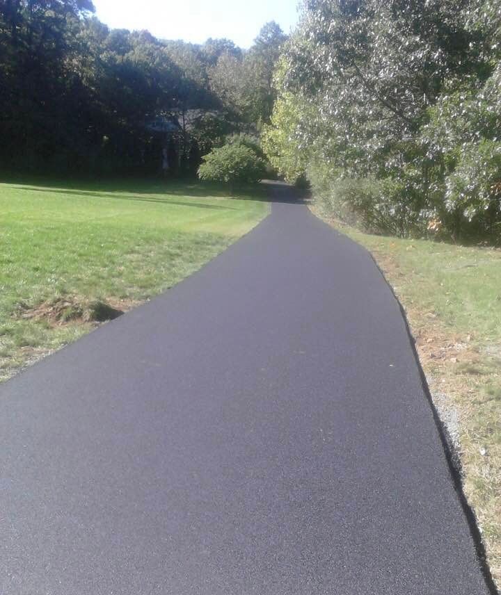 A freshly paved, dark asphalt driveway curves through a grassy, tree-lined residential landscape on a sunny day.