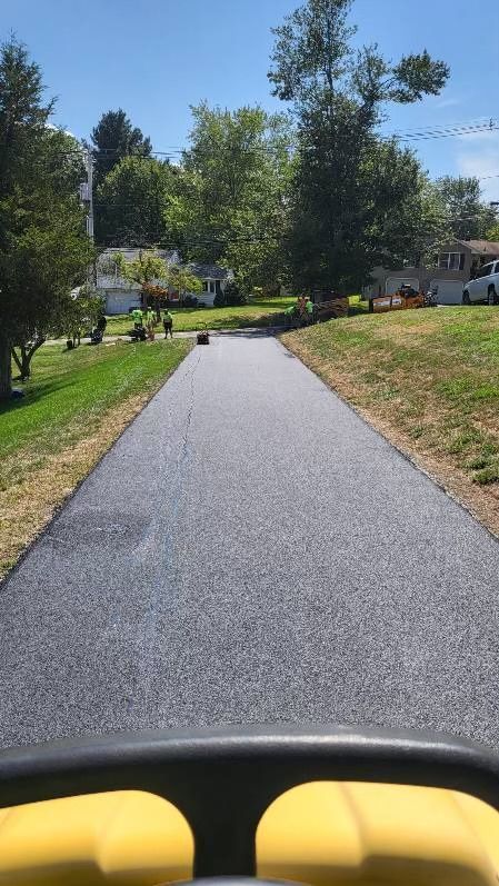 A view from the driver's seat of a yellow vehicle moving down a newly paved asphalt driveway on a sunny day.
