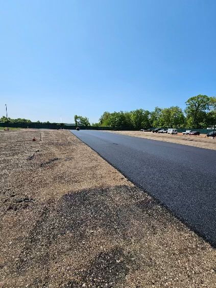 A newly paved, dark asphalt road stretches through a sunny field under a clear blue sky, next to a line of trees.