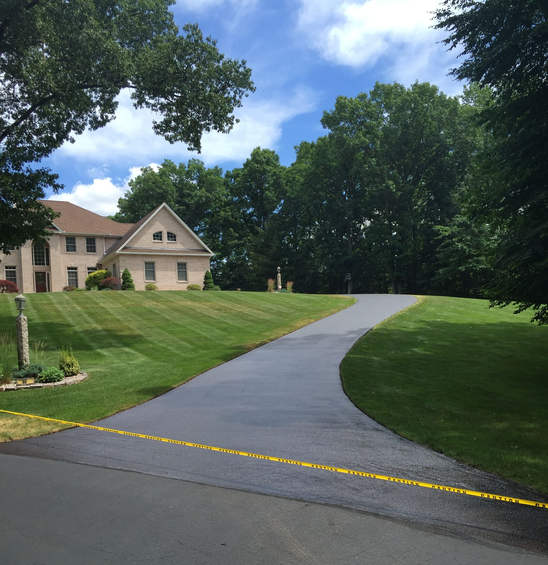 A newly paved dark asphalt driveway leads up a grassy slope toward a large multi-story tan house nestled in dense trees.