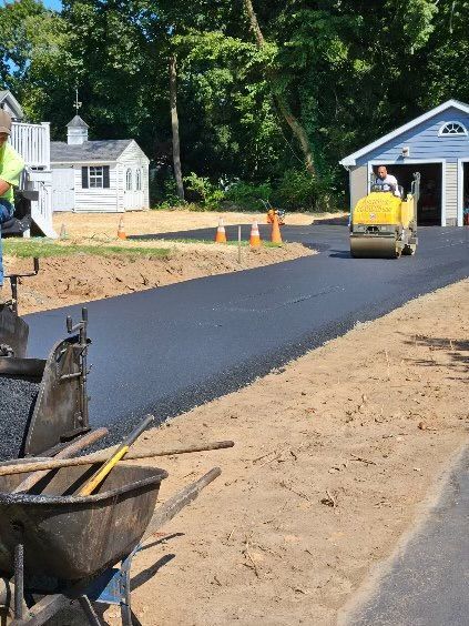 A construction worker operates a yellow roller to smooth fresh asphalt on a residential driveway near a house.