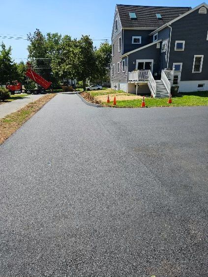 A newly paved asphalt driveway leads toward a gray house with a white deck, with construction cones and trees nearby.