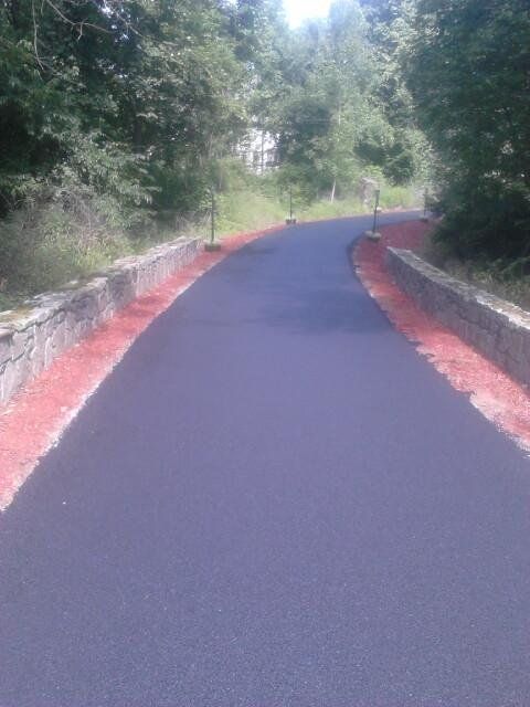 A freshly paved, dark asphalt path bordered by reddish-brown mulch and low stone walls, leading into a green forest.