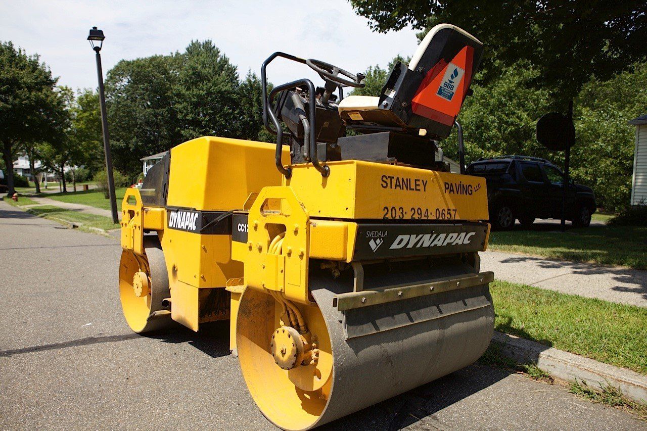 A yellow Dynapac construction roller parked on an asphalt street with a grassy yard and trees in the background.