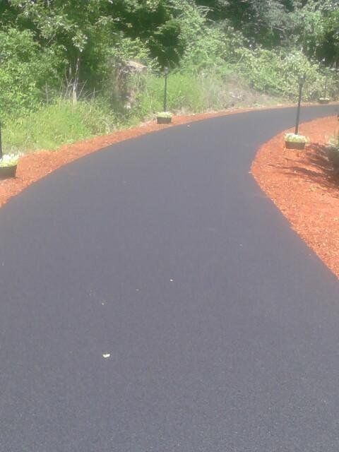 A freshly paved, curved asphalt driveway lined with red mulch and small, decorative lights in a wooded setting.