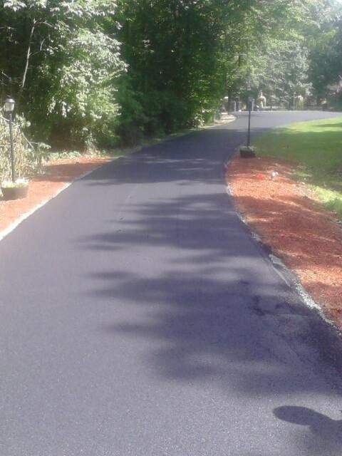 A freshly paved black asphalt driveway curves between lush green trees and beds of red mulch.