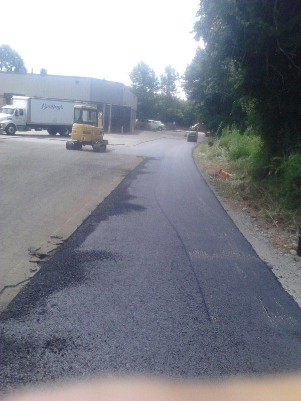 A freshly paved asphalt path leads toward a construction site with a yellow excavator and a parked white semi-truck.