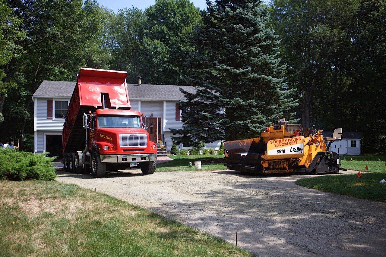A red dump truck and a yellow paving machine sit on a gravel driveway in front of a house surrounded by trees.