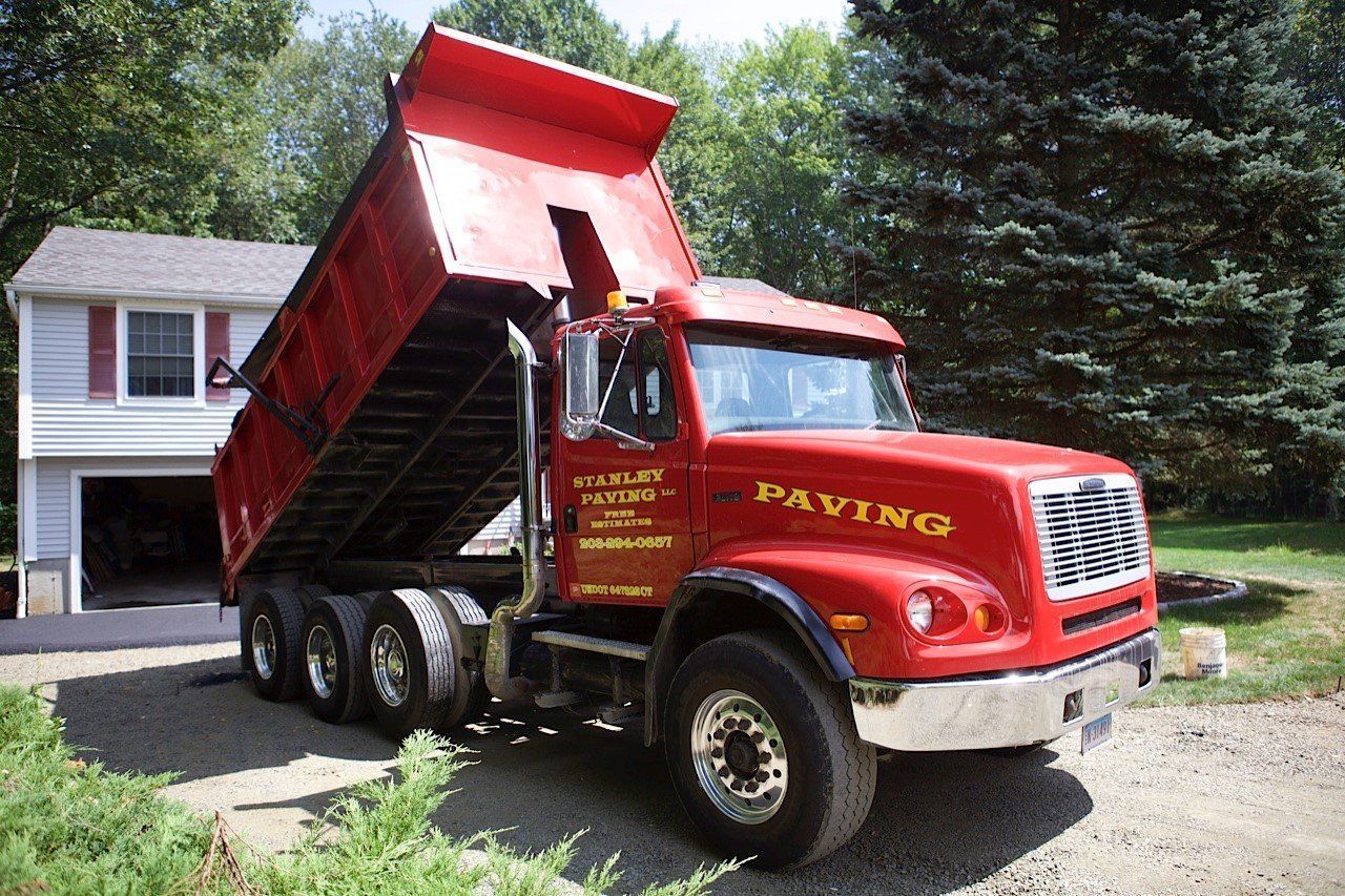 A red dump truck with its bed raised, parked on a gravel driveway in front of a house.