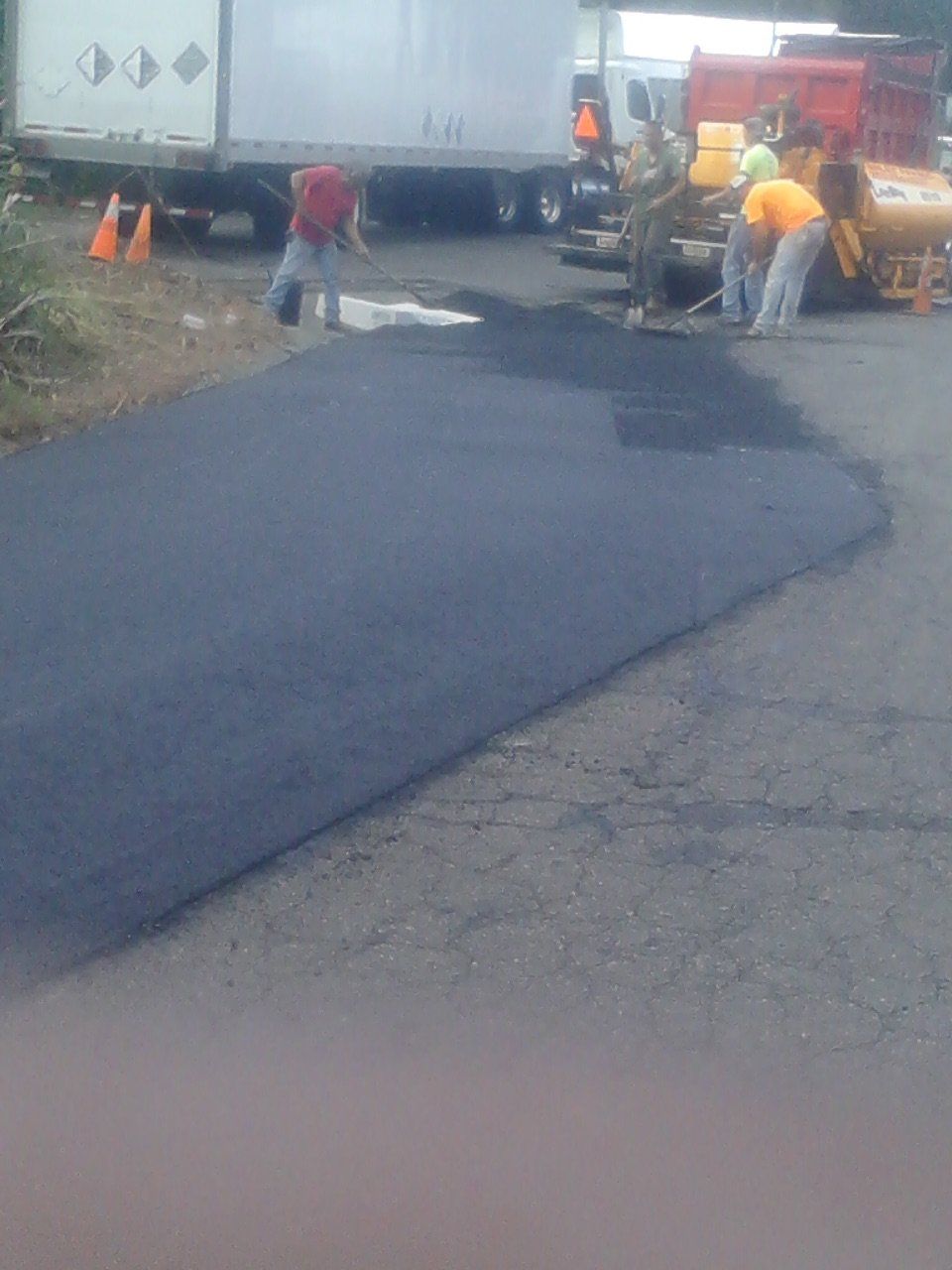 A construction crew works on a patch of freshly laid black asphalt on a paved road next to a large white truck.