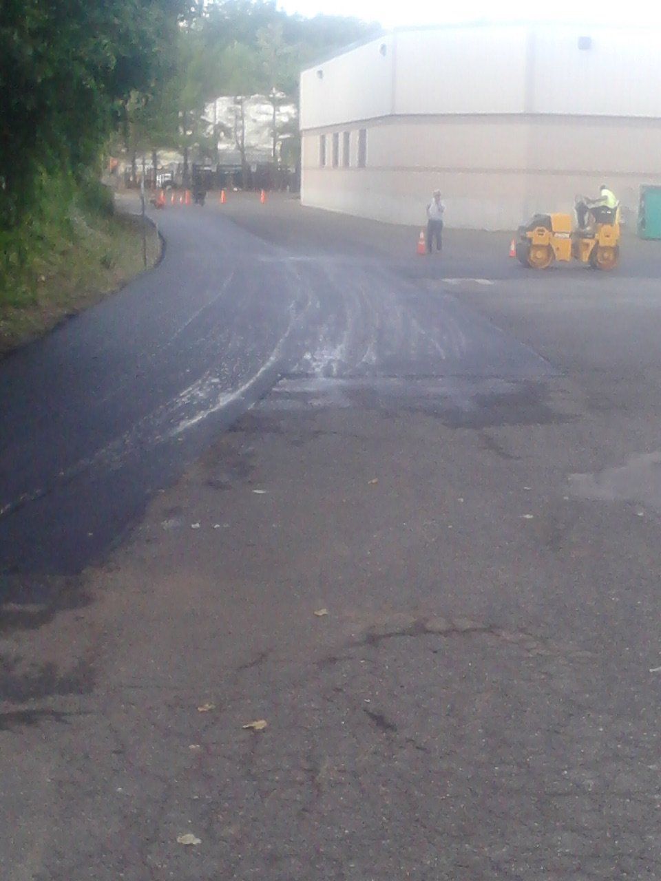 A worker operates a yellow steamroller on a newly paved black asphalt road next to a large building.