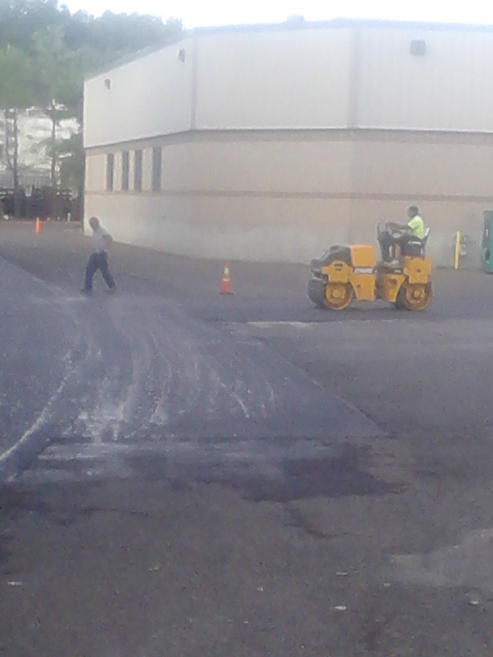 A construction worker operates a yellow asphalt roller near a building while a pedestrian walks across the lot.