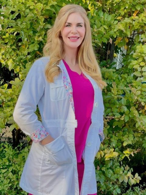 Krista Smith in a lab coat and pink scrubs is standing in front of trees.