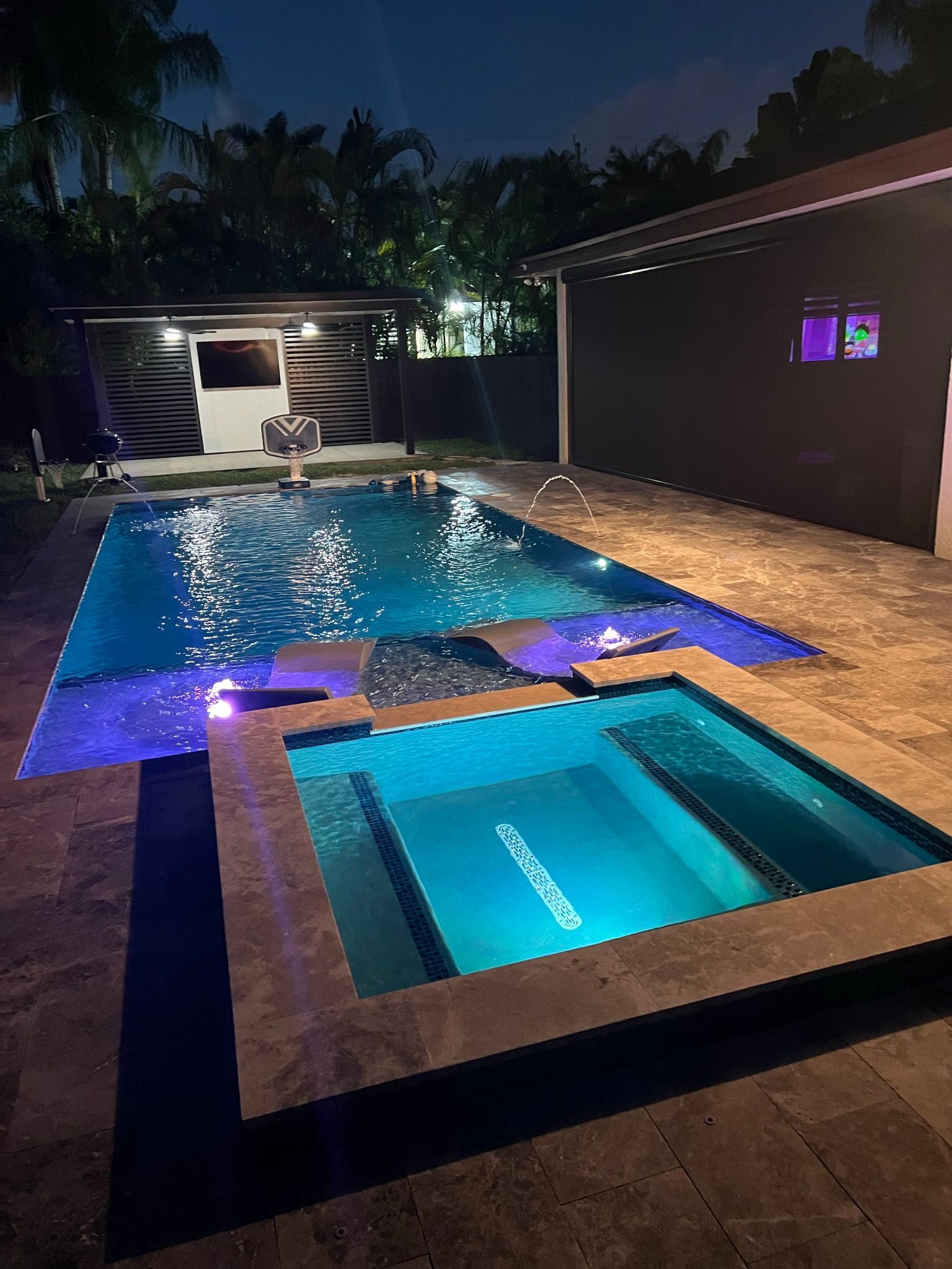 A large swimming pool and hot tub are lit up at night.
