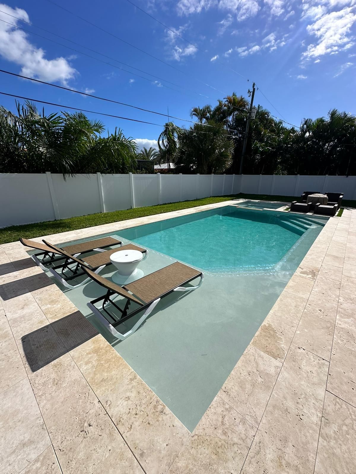 A backyard pool with lounge chairs, surrounded by light-colored stone. Blue sky above, with trees and white fence in background.
