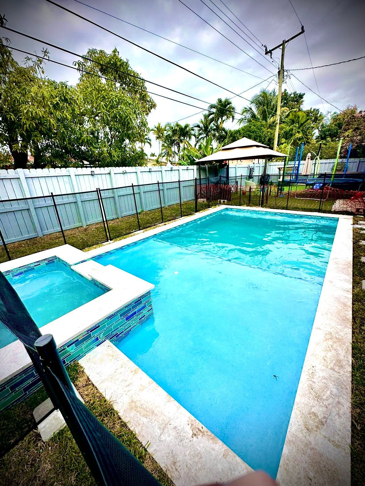 Pool with attached hot tub, enclosed by a fence, with a gazebo in the background under a cloudy sky.