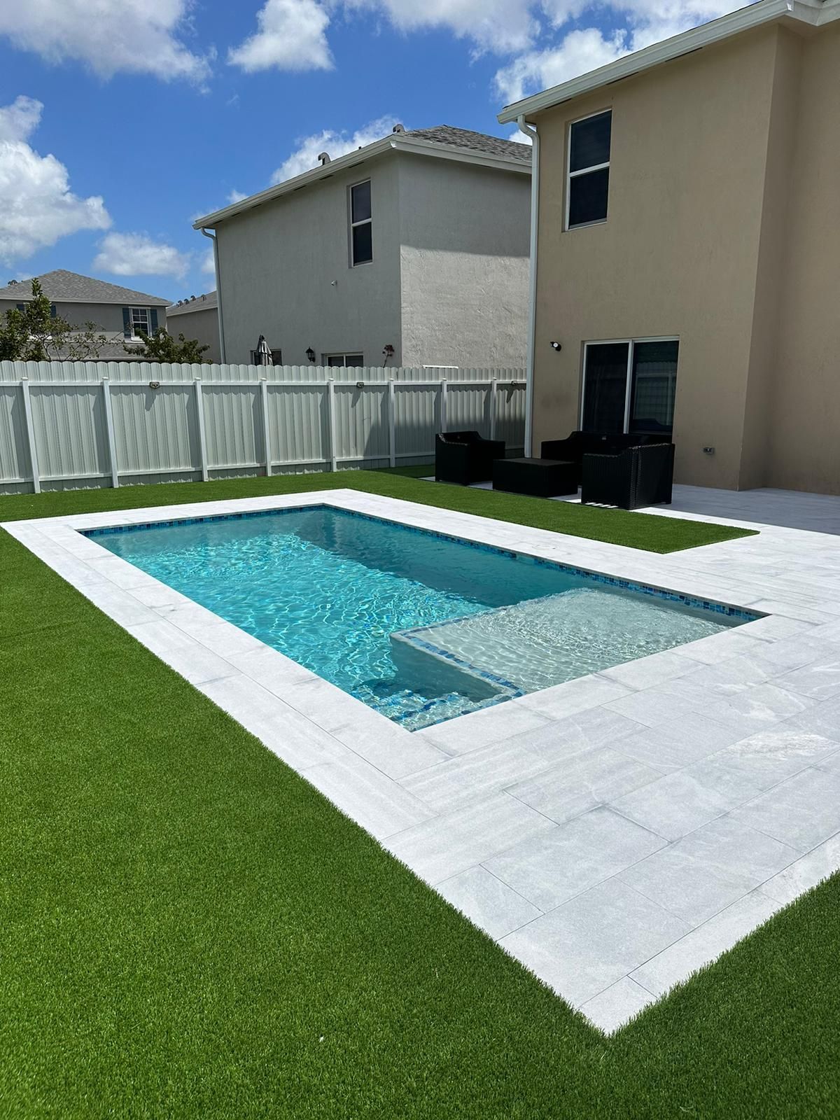 Backyard with a rectangular pool, white pavers, and artificial green grass. Two-story houses and a white fence are in the background.