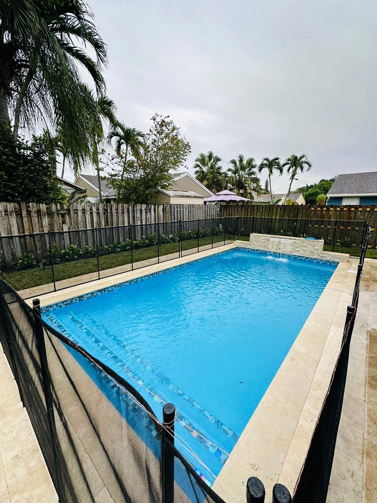 Rectangular swimming pool with blue water, surrounded by light-colored stone. Black safety fence borders the pool, set in a backyard with trees and a wooden fence.