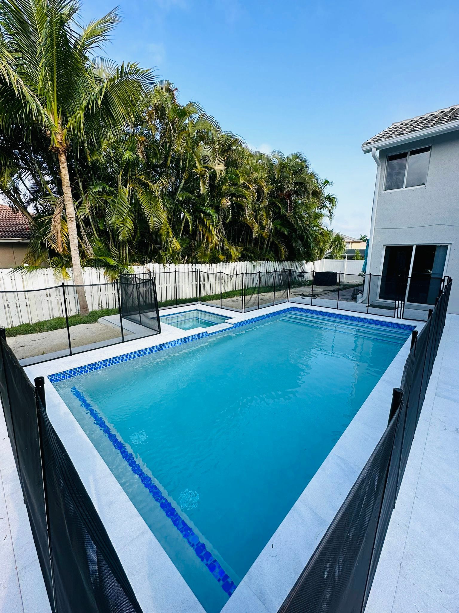 A backyard with a rectangular pool, hot tub, and a two-story house. A black fence surrounds the pool area.