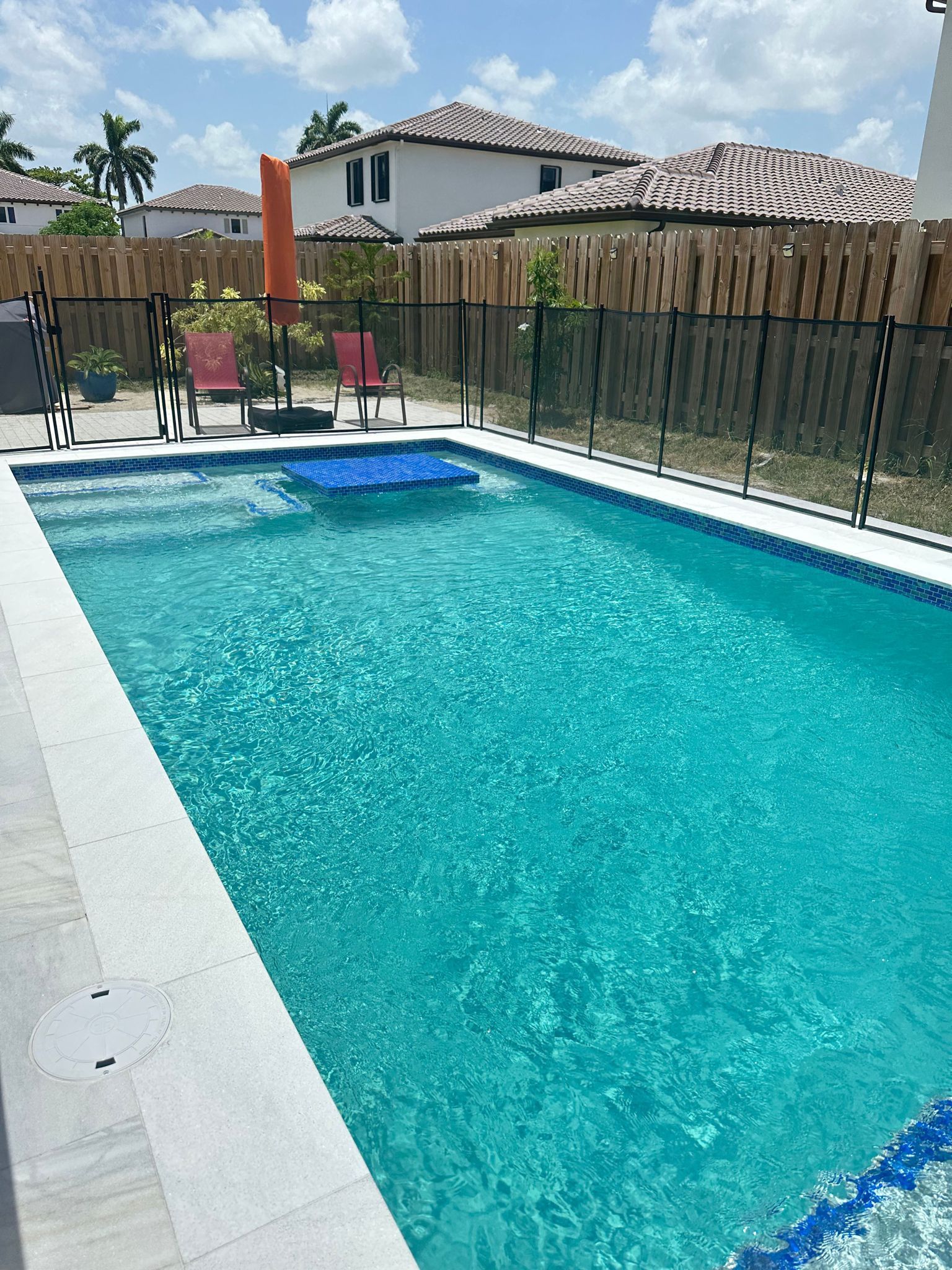 A rectangular turquoise swimming pool surrounded by a light-colored deck and a black fence. Two red chairs sit near an orange umbrella.