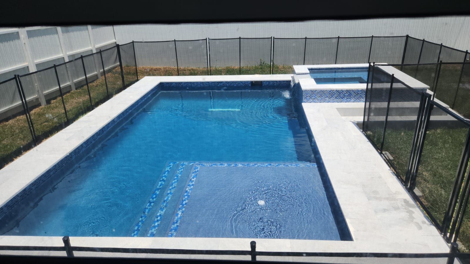 A large swimming pool surrounded by a fence and a hot tub.