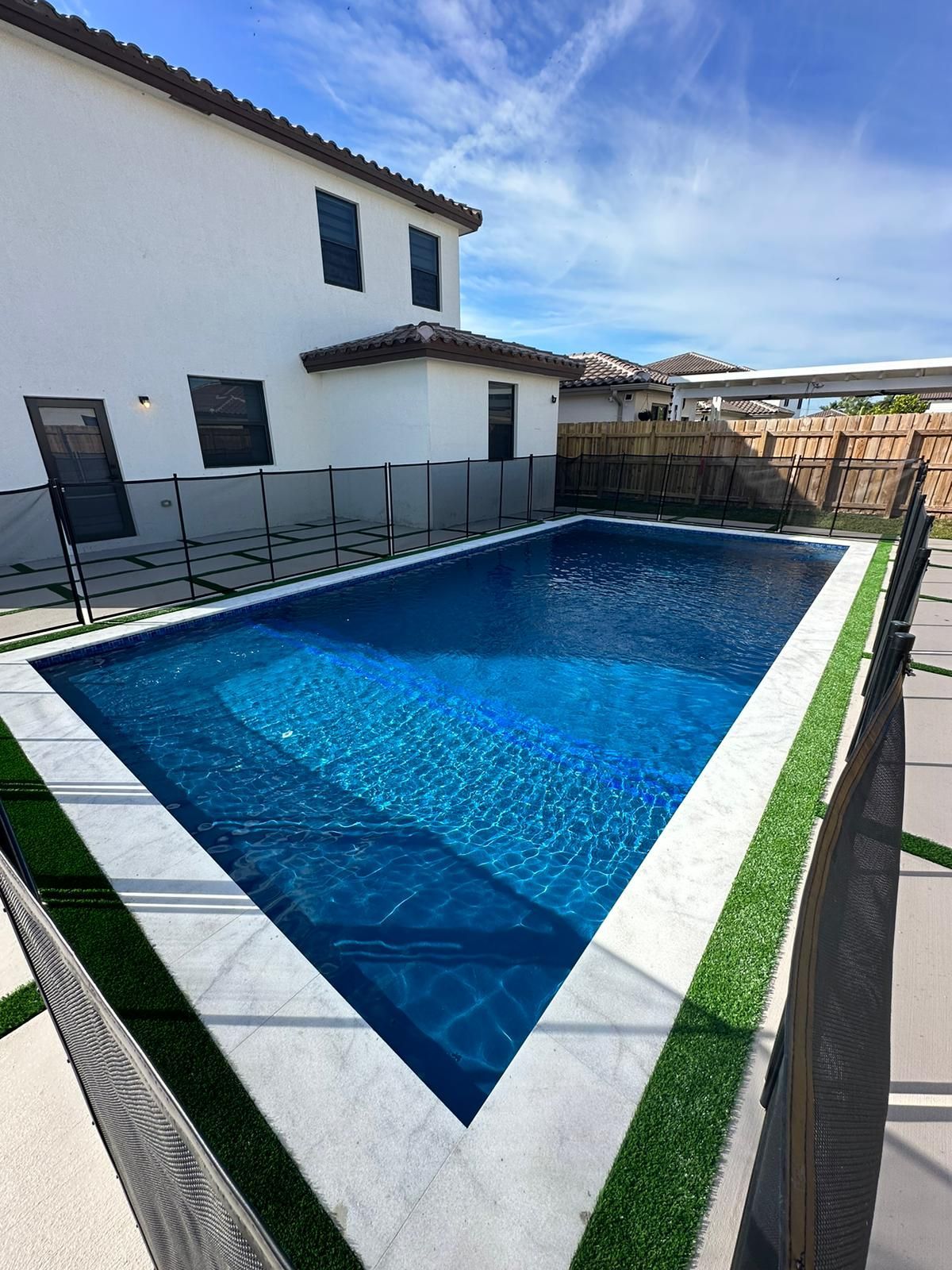 A large swimming pool is surrounded by a fence and grass in front of a house.