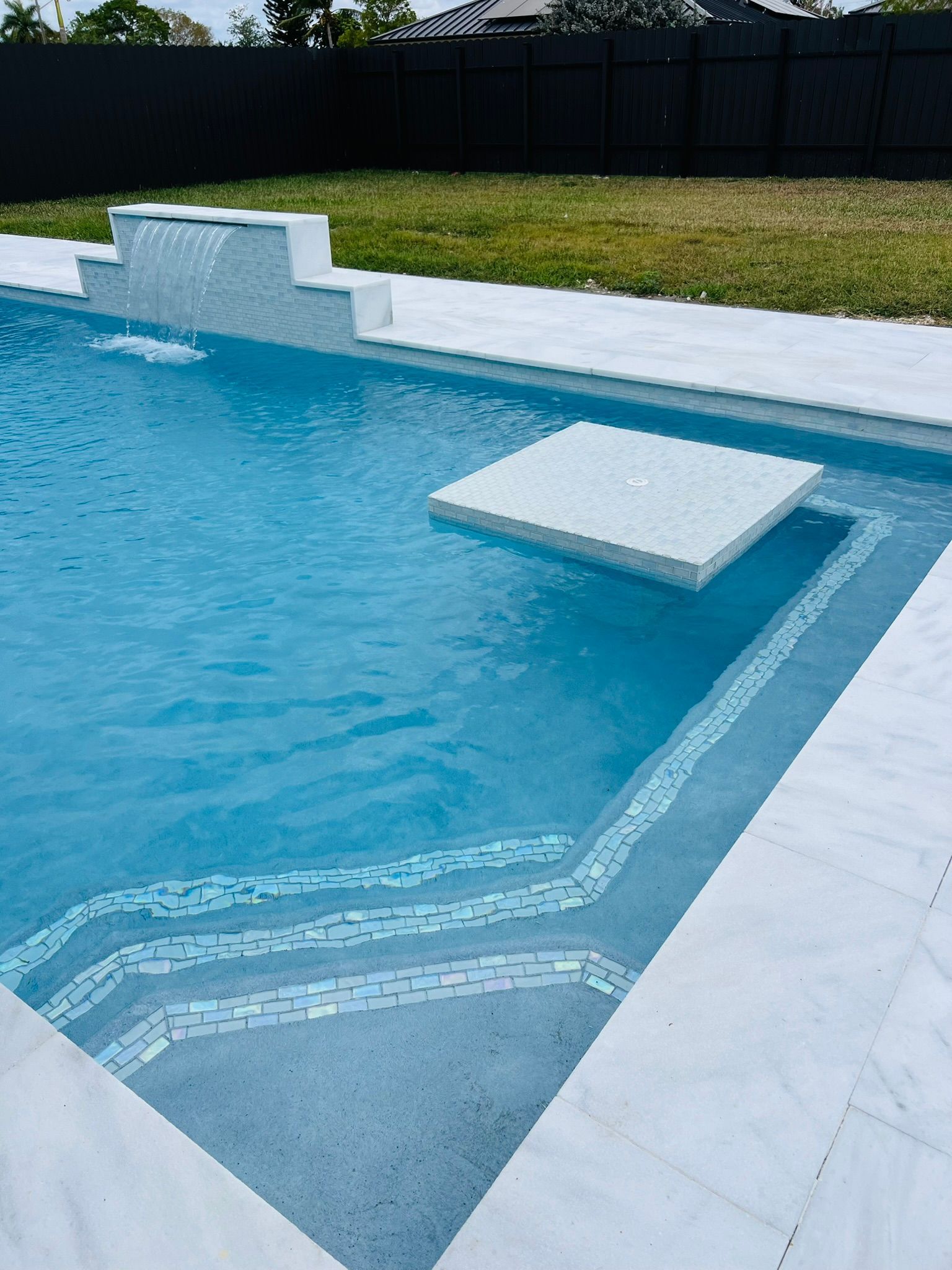 A large swimming pool with steps and a waterfall in the backyard.