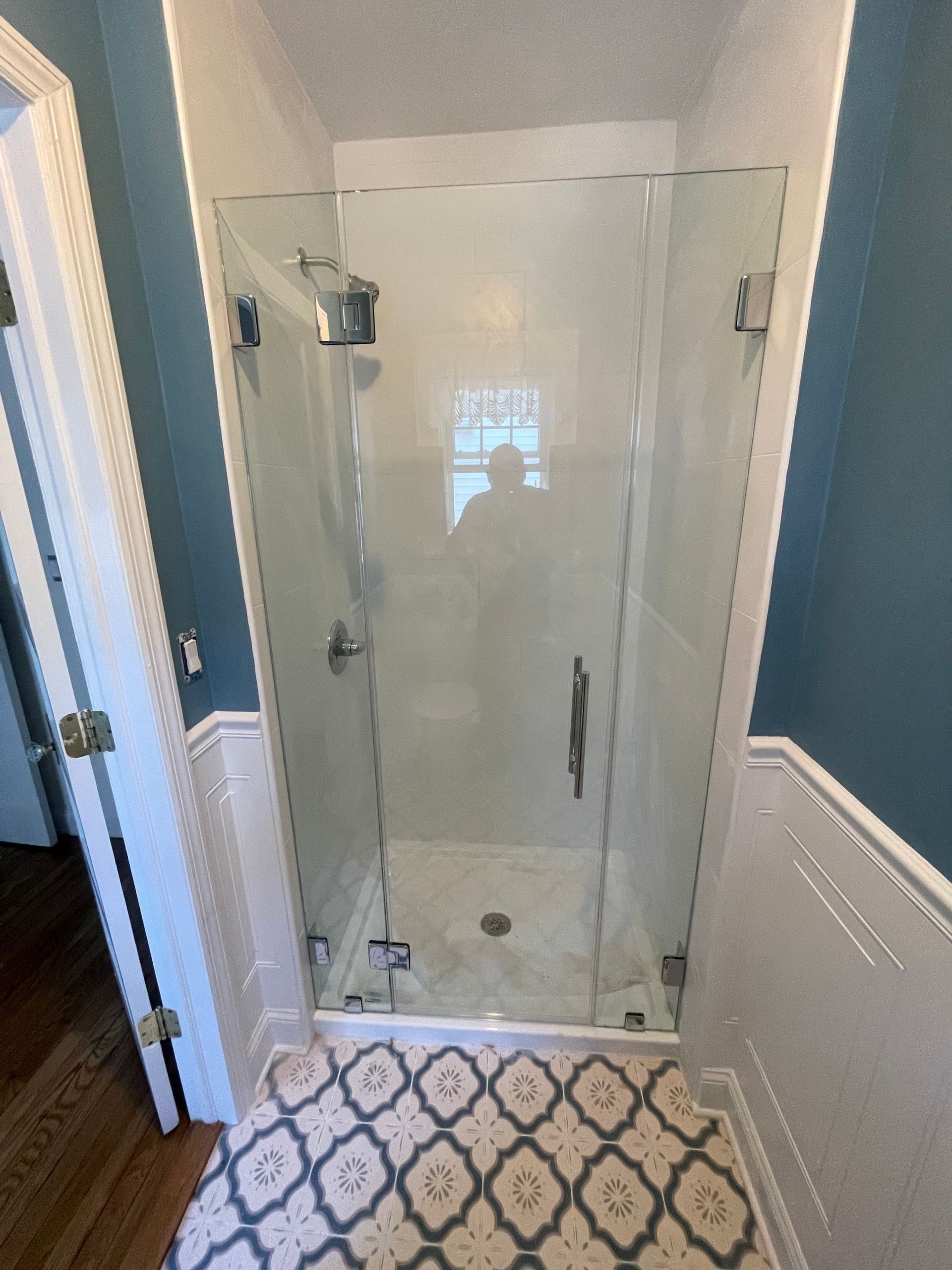 Bathroom with glass shower enclosure, blue walls, white wainscoting, and patterned tile floor.