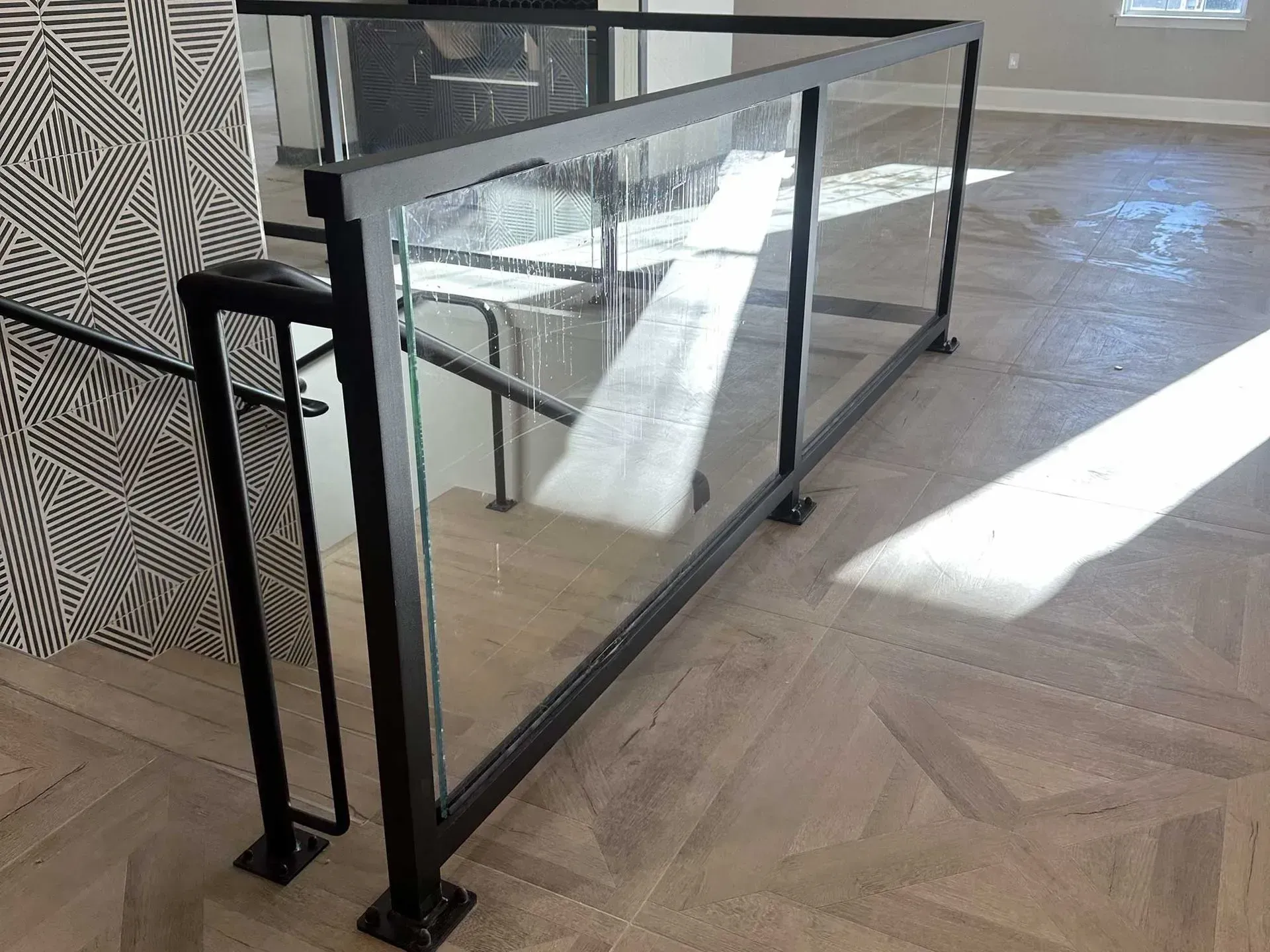 Black metal staircase railing with glass panels installed on a patterned wood floor.