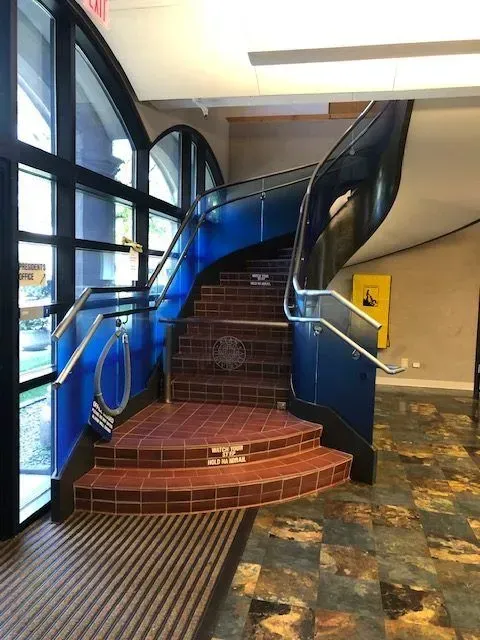 A tiled staircase curves upward between a glass wall and a dark blue railing in a building lobby.