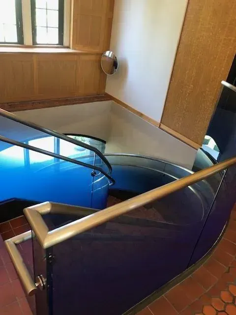 A spiral staircase with metallic railings and blue glass side panels inside a building with a tile floor and wooden wall.