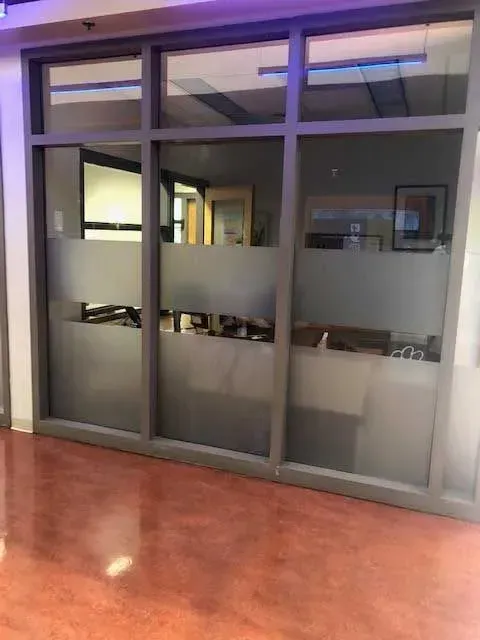 A glass office partition with grey metal frames and horizontal frosted bands, set against a polished reddish-brown floor.