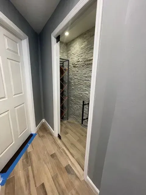 A hallway with wood-look flooring and gray walls leads to a wine closet featuring stone-tiled walls and metal racks.