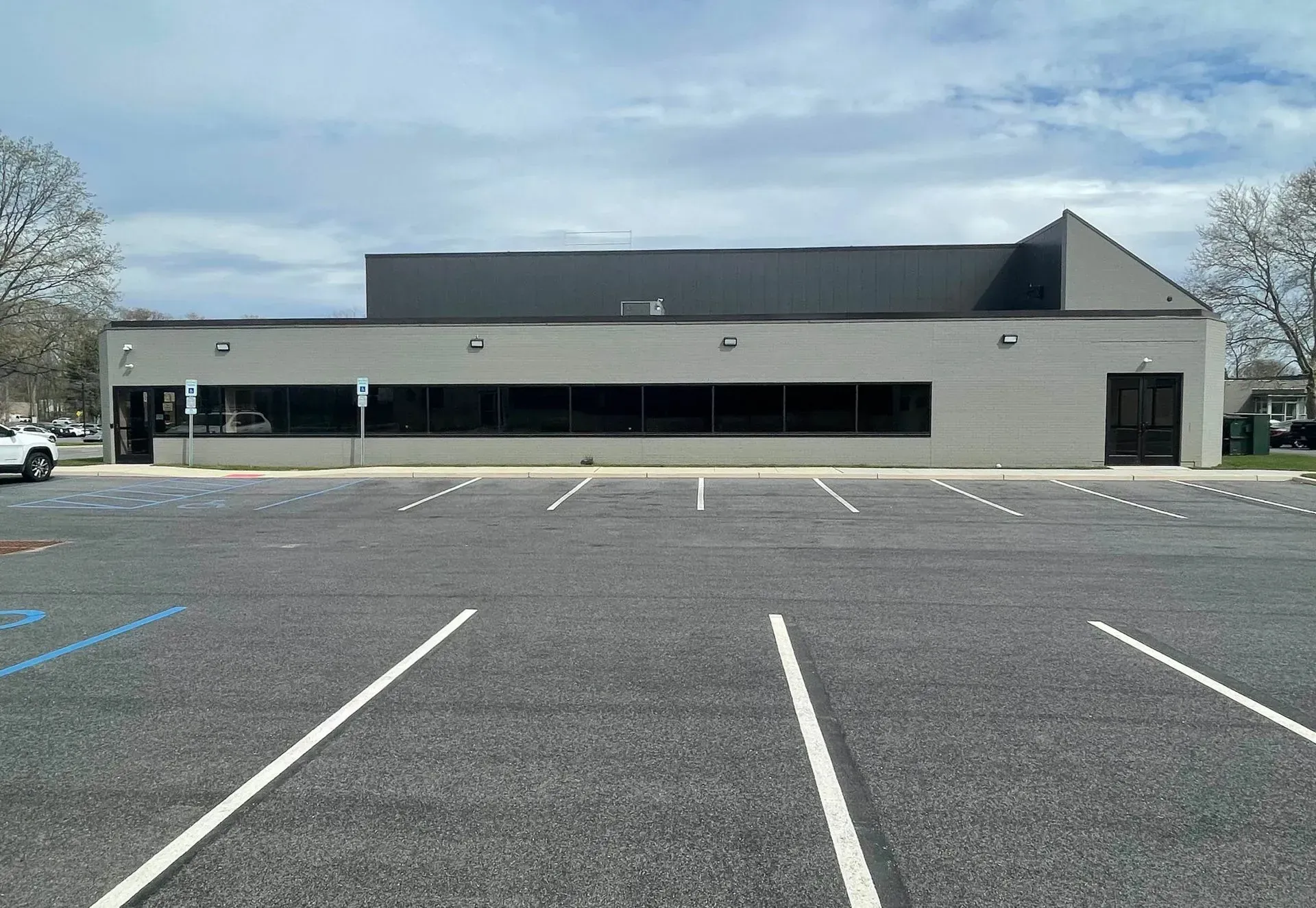 A grey, single-story commercial building with a long band of dark windows and a large, empty asphalt parking lot.