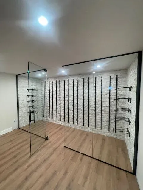 A modern wine cellar with glass walls, light wood flooring, a textured white stone wall, and black metal bottle racks.