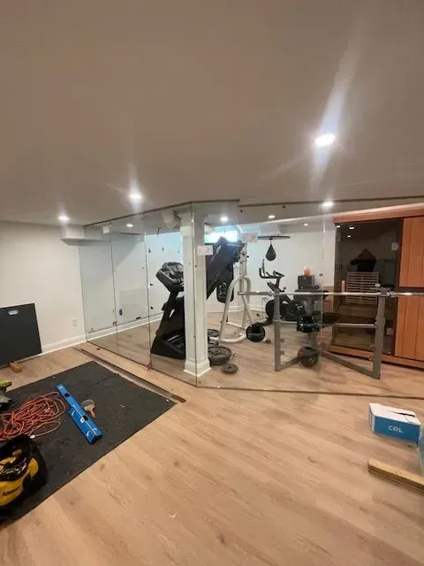 A basement home gym with light wood floors, exercise equipment, a sauna, and glass-partitioned walls under recessed lights.