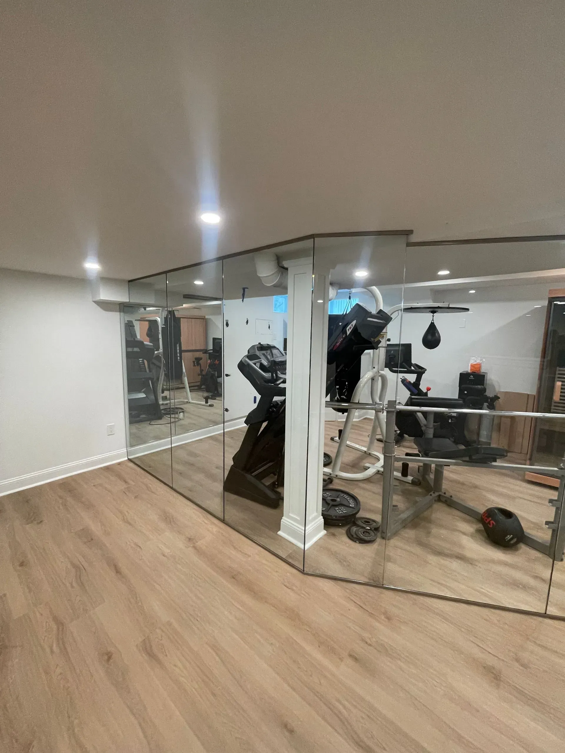 A home gym area featuring wood-look flooring, a white structural pillar, and a large wall mirror reflecting workout equipment.