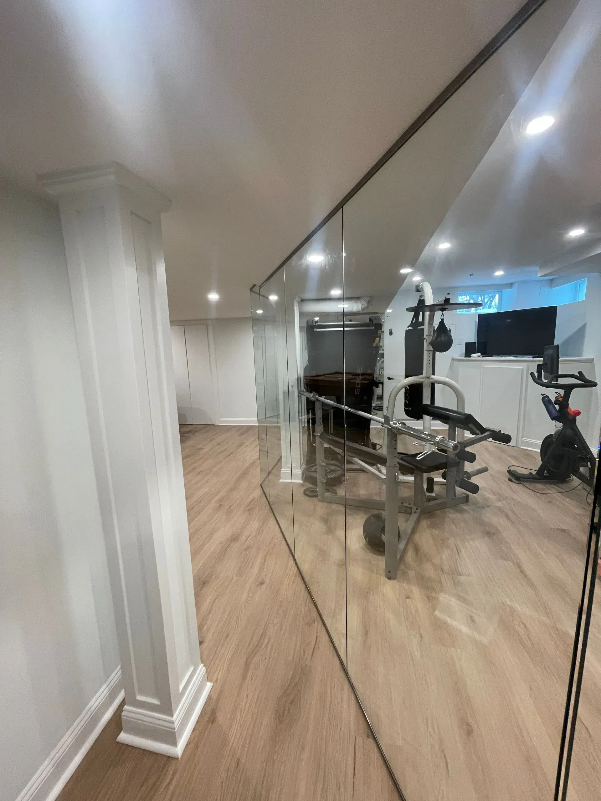 A home gym with light wood floors, mirrored walls, a weight bench, and a stationary bike in a bright, modern basement.